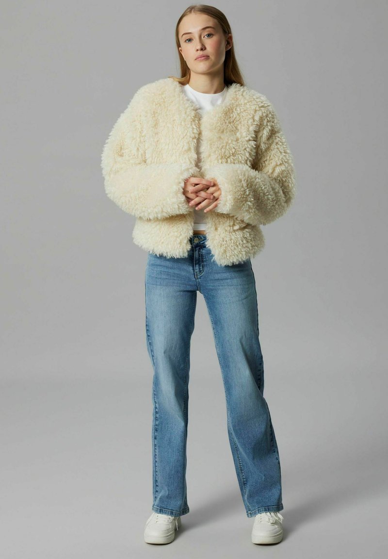 Neppelsen jassen in crème met een boxy silhouet, brede mouwen en een ronde halslijn, gecombineerd met lichtblauwe flared jeans en witte sneakers.