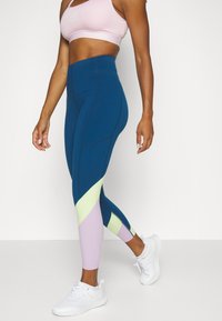 Žena v růžovém sportovním podprsence, legínách s barevnými bloky v modré, světle zelené a levandulové barvě, a bílých sportovních botách, stojí s jednou nohou vpřed.