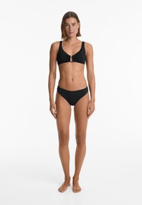 Il set di bikini nero presenta un top arricciato con un dettaglio a chiave sul davanti e uno slip alto coordinato. Tessuto liscio, cuciture minime.