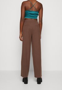 Pantalones anchos marrones con bolsillos traseros, combinados con un top corto de satén en tono verde azulado con tirantes entrelazados. Zapatos planos complementan el conjunto.