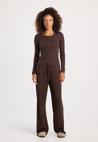 Bruine lange mouwen top met knoopdetails en bijpassende losse broek. Zachte stof met een gladde textuur, aangevuld met beige slippers.