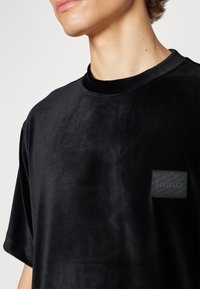 Gros plan d'une personne portant un T-shirt en velours côtelé noir avec une petite étiquette noire "HUGO" sur la poitrine gauche.