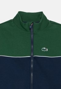 Jachetă cu fermoar din bumbac în verde închis și albastru marin, având un guler înalt, o linie de accent albă și un mic logo brodat cu crocodil.