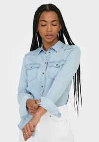 Camisa de denim azul claro con cierre de botones, dos bolsillos en el pecho y mangas enrolladas. Textura suave y diseño de cuello clásico.