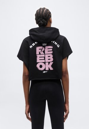 Naine punutud kukliga, seljas must kärbitud kapuutsiga dressipluus roosa "REEBOK" tekstiga ja mustad retuusid, seisab seljaga kaamerasse tavalise tausta ees.