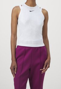 Personne portant un haut sans manches blanc Nike et un pantalon violet taille haute, debout les bras détendus le long du corps.