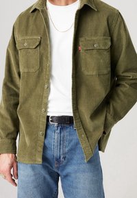 Camicia in corduroy verde oliva con due tasche sul petto, chiusura a bottoni e colletto, abbinata a una maglietta bianca e a jeans in denim blu.
