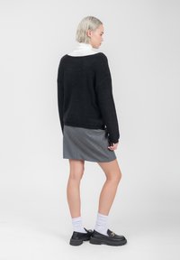 Zwarte gebreide trui met een losse pasvorm, gecombineerd met een grijze rok met witte stippenpatronen. Model draagt zwarte instappers en witte sokken.