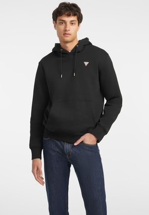 Schwarzer Hoodie mit Kordelzügen und einem kleinen dreieckigen Logo auf der Brust, kombiniert mit blauen Jeans im Denim-Stil. Schlichte Textur, lockere Passform.