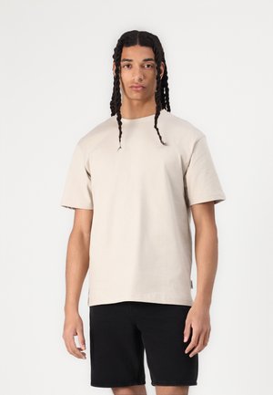 Beige bomulds t-shirt med afslappet pasform, korte ærmer og rund halsudskæring, parret med sorte shorts, præsenteret på en neutral baggrund.