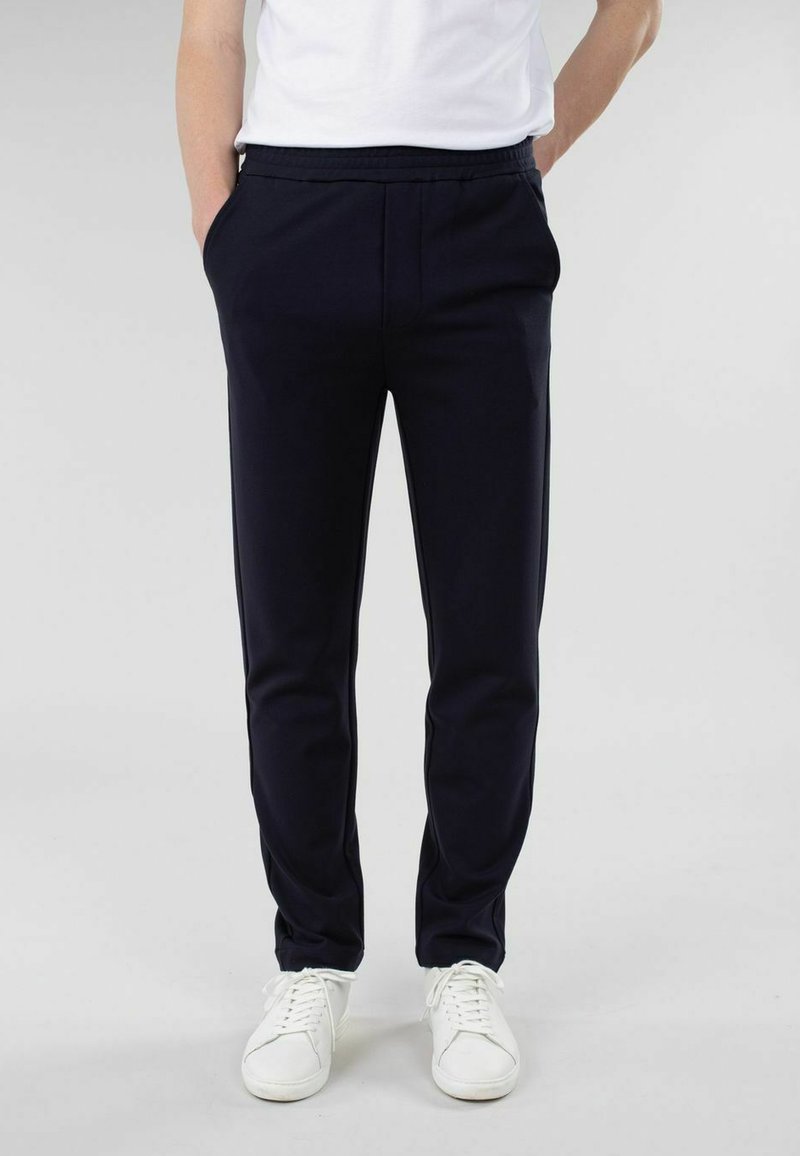 Mørkeblå, slim-fit bukser med et glat stof, elastisk talje, sidelommer og et avsmalnende ben-design. Kombinert med hvite joggesko.