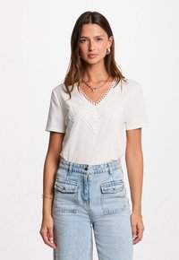 Mujer con cabello largo castaño, vestida con una blusa blanca de cuello en V con detalle de encaje y jeans de tiro alto de color azul claro con bolsillos delanteros.