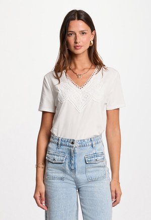 Donna con lunghi capelli castani che indossa un top bianco con scollo a V e dettagli in pizzo, abbinato a jeans azzurri a vita alta con tasche frontali.