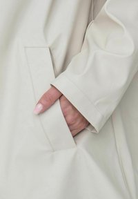 Manteau beige clair, imperméable, avec une texture lisse. Doté d'une poche latérale et d'une manche légèrement retournée, mettant en valeur des détails de couture précis.