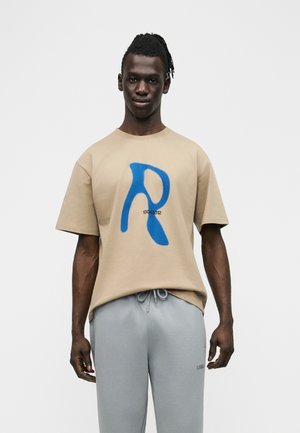 Vyro su tamsia oda ir surištais plaukais, vilkinčio smėlio spalvos marškinėlį su didžiule mėlyna raide „R“ ir šviesiai pilkus sportinius kelnes, stovinčio prieš paprastą foną.