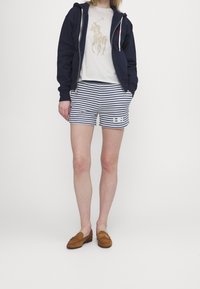 Marineblå zip-up hoodie över en krämfärgad skjorta med polo-spelargrafik, randiga shorts och bruna mockaloafers.