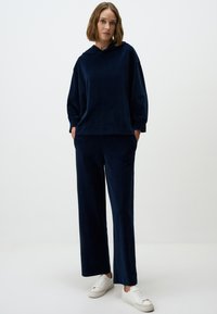 Sweat à capuche en velours bleu marine avec une poche avant et des manches longues, associé à un pantalon large assorti, complété par des baskets blanches.
