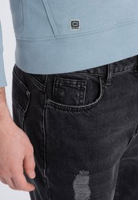 Sudadera azul clara con dobladillo y puños acanalados, con una pequeña etiqueta de logo. Combinada con jeans de mezclilla negra desgastados. Mano posicionada en la cintura.