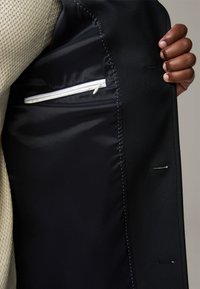 Zwart maatpakjack met een gladde satijnen voering, voorzien van een witte ritszak en een extra klein zakje; gestructureerd ontwerp.