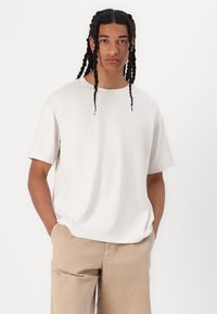 Camiseta blanca de manga corta con un ajuste relajado y escote redondo, mostrada en una persona que lleva pantalones beige. Tejido de algodón suave.
