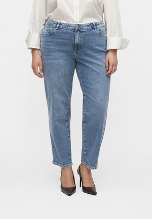 Femme portant un jean droit bleu clair, une chemise blanche à manches longues et des chaussures à talons hauts pointues noires, debout devant un fond blanc.