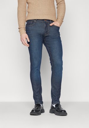 Jeans en denim slim-fit bleu foncé avec cinq poches et des coutures jaunes contrastantes. Portés avec un pull à côtes beige et des mocassins noirs.
