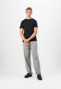 Ung mann står med hendene foldet, iført svart t-skjorte, lysegrå jeans og svarte sko mot en helt hvit bakgrunn.