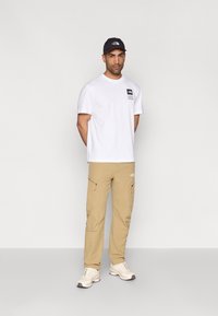 Λευκό κοντομάνικο μπλουζάκι με λογότυπο, χακί cargo παντελόνι με τσέπες και γκρι καπέλο. Λευκά αθλητικά παπούτσια με γκρι λεπτομέρειες. Απλό φόντο.