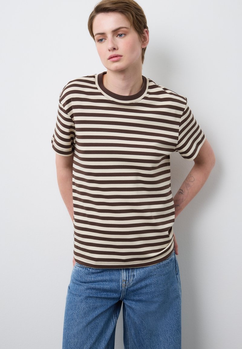 Persona con cabello corto, vistiendo una camisa a rayas marrón y beige y vaqueros azules sueltos, de pie con las manos detrás de la espalda contra una pared blanca.