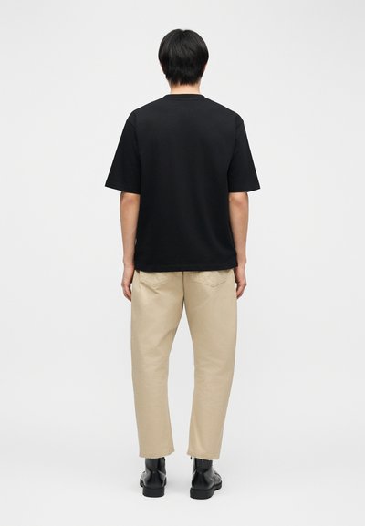 Άνδρας που στέκεται με την πλάτη γυρισμένη, φορώντας μια απλή μαύρη κοντομάνικη μπλούζα, μπεζ παντελόνι και μαύρα μποτάκια αστραγάλου σε λευκό φόντο.