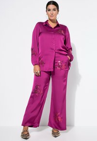 Magenta satijnen shirt en wijde broekset, met bloemenborduursel. Het shirt heeft een knoopsluiting; de broek heeft zijzakken en een gladde textuur.
