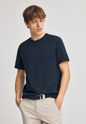 Marine crew-neck t-shirt lavet af blødt stof, med korte ærmer og et lille broderet logo på brystet, sat sammen med beige bukser og et bælte.