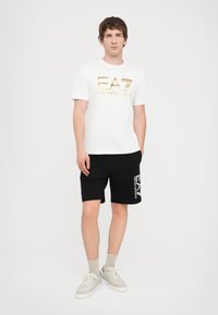 Hvit t-skjorte med gull EA7-logo, sammen med svarte shorts med samme logo. Fotwear er hvite med grå detaljer.