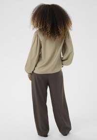 Olívazöld blúz ráncolt ujjal, laza szabású barna nadrággal párosítva. Simma textúra, egyszerű dizájn. Egyszerű háttér.