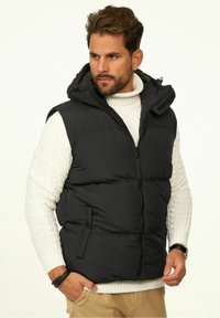 Black puffer vest with a hood, zippered front, and side pockets, worn over a white cable-knit sweater. Features a smooth, insulated texture.
