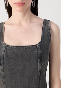 Et nærbilde av en kvinnes overkropp iført en ermeløs grå denimkjole, med minimalistiske sølvøredobber, mot en nøytral bakgrunn.