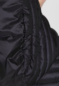 Detailní záběr na černou prošívanou látku s lesklou, mírně zmuchlanou strukturou a s vertikálními prošitými švy.
