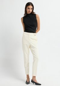 Jonge vrouw staat tegen een witte achtergrond en draagt een mouwloze zwarte top, crèmekleurige high-waisted broek met riemdetails en zwarte hakken.