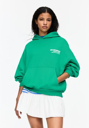 Green hooded sweatshirt in soft fabric, featuring a front pocket and white text logo. Paired with a white pleated skirt at the hem.