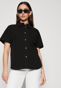 Camicia nera a maniche corte con colletto e bottoni in argento, realizzata in tessuto di denim. Abbinata a pantaloni bianchi e occhiali da sole scuri.
