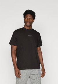 T-shirt preta de algodão com mangas curtas e gola redonda, apresentando um pequeno logotipo em texto branco no peito. Calças de moletom cinzas visíveis.