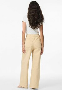Pantalons larges beige clair en coton doux avec poches arrière. Le mannequin porte un t-shirt blanc ajusté, mettant en valeur le style décontracté des pantalons.