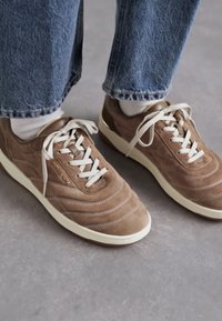 Sneaker in suede marrone con lacci bianchi, caratterizzate da una superficie texturizzata, cuciture a strati e suola in gomma bianca. Indossate con jeans blu.