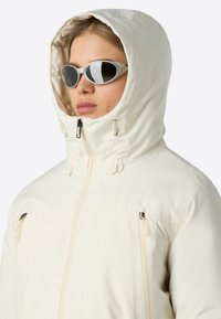 En person iklädd en krämfärgad huva-jacka som är zipad till toppen och vita ovala solglasögon, vänd något åt vänster.