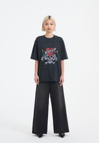 Schwarzes Grafik-T-Shirt mit einem Totenkopf- und Herzdesign sowie Text, kombiniert mit weit geschnittenen schwarzen Jeans. Durchgehend einfarbig.