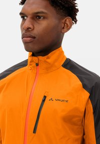 Giacca leggera arancione e nera con zip e colletto alto, pannelli a contrasto, zip frontale e piccola tasca sul lato sinistro del petto. Logo Vaude sulla parte frontale.