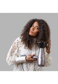 Στεγνωτήρας μαλλιών με μεταλλικό ασημί σώμα, μαύρη μ�ύτη και κυκλική είσοδο αέρα. Το μοντέλο τον κρατάει φορώντας μια άσπρη μπλούζα με πολύπλοκο σχέδιο.