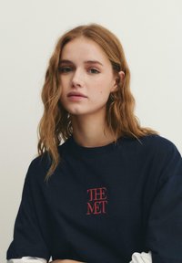 Junge Frau mit welligem hellbraunem Haar, die ein dunkelblaues Hemd mit rotem Stickschriftzug "THE MET" vor einem einfarbigen hellen Hintergrund trägt.