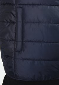 Laivastonsininen toppaliivi, jossa vaakasuora tikkaus ja sivutasku. Sileä, kiiltävä viimeistely kevyellä, eristävällä vaikutelmalla.