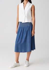 Vit ärmlös kavaj med en knapp framtill, parat med en flödande midi-skjol i blå denim med volangmidja och vita sneakers.